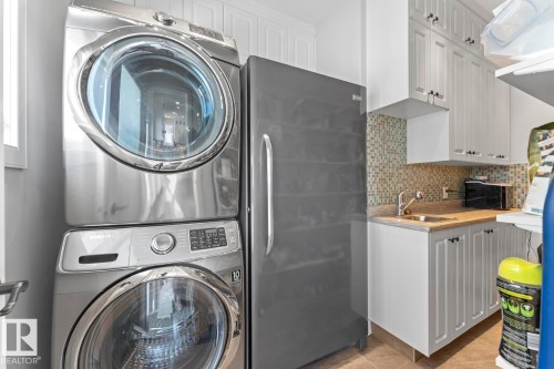This utility area features a stackable washer and dryer, a stainless steel refrigerator, white cabinetry with dark hardware, a sink, and a tiled backsplash - 1513 Horseshoe Bay Estates, Cold Lake, AB - Indoor Photo Showing Laundry Room