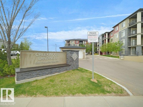 Entry sign for Creekwood Landing II featuring stacked stone accents, green landscaping, and a paved street leading to multi-story buildings with balconies - 216 5390 Chappelle Road, Edmonton, AB - Outdoor With Balcony