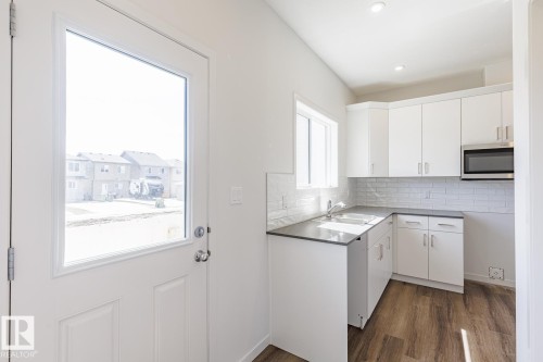 Kitchen featuring white cabinetry, subway tile backsplash, dark countertops, a stainless steel microwave, and wood-finish flooring - 3613 Triomphe Boulevard N, Beaumont, AB - Indoor Photo Showing Kitchen
