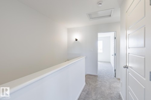 Carpeted hallway featuring a white half-wall, modern interior doors with silver hardware, a ceiling-mounted smoke detector, and an overhead attic access panel - 3613 Triomphe Boulevard N, Beaumont, AB - Indoor Photo Showing Other Room