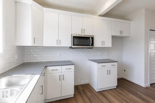 Kitchen featuring white cabinetry with brushed nickel hardware, a stainless steel microwave, gray countertops, a subway tile backsplash, and wood-finish flooring - 3613 Triomphe Boulevard N, Beaumont, AB - Indoor Photo Showing Kitchen With Double Sink