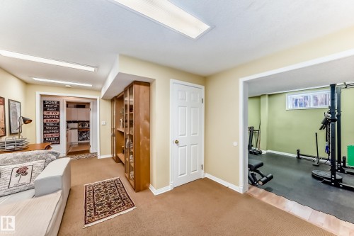 Carpeted recreation area featuring a built-in display cabinet with glass doors, a white paneled door, and an open doorway to an exercise room with rubber flooring - 11115 44 Street, Edmonton, AB - Indoor