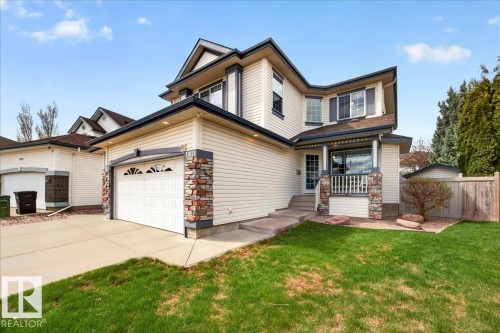 Cream-colored siding exterior featuring a covered front porch with stone accents, an attached garage with decorative windows, a concrete driveway, and a front lawn with established landscaping - 11824 13 Avenue, Edmonton, AB - Outdoor
