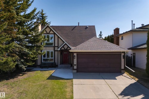 Tudor-style exterior featuring stucco and exposed timber framing, a red entry door, a two-car garage with a brown door, mature evergreen landscaping, and a concrete driveway - 15251 88A Street, Edmonton, AB - Outdoor