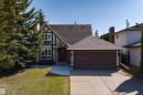 Tudor-style exterior featuring stucco and exposed timber framing, a red entry door, a two-car garage with a brown door, mature evergreen landscaping, and a concrete driveway - 15251 88A Street, Edmonton, AB  - Outdoor 