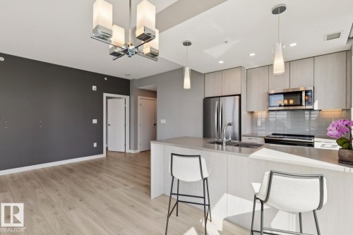 Open-concept kitchen featuring light wood-finish flooring, a contemporary chandelier, stainless steel appliances, a light-toned countertop, and modern cabinetry - #1601 10180 103 St Nw, Edmonton, AB - Indoor Photo Showing Kitchen With Upgraded Kitchen