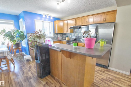 Kitchen featuring wood-finish flooring, a central island with a countertop, wood cabinetry, a mosaic tile backsplash, and stainless steel appliances - 14820 140 Street, Edmonton, AB - Indoor Photo Showing Kitchen
