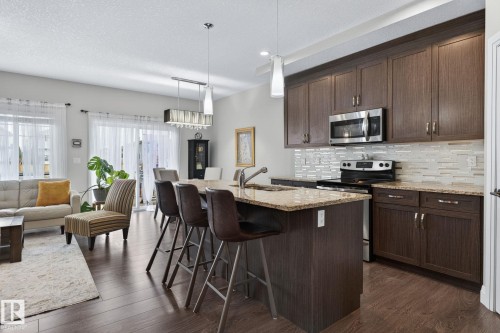 Open-concept kitchen featuring dark wood-finish cabinetry, stainless steel appliances, a light-toned granite countertop, and a mosaic tile backsplash - 83 Sandalwood Place, Leduc, AB - Indoor Photo Showing Kitchen With Upgraded Kitchen