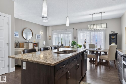Kitchen island featuring a granite countertop with integrated double basin sink, dark wood-finish cabinetry, and pendant lighting - 83 Sandalwood Place, Leduc, AB - Indoor Photo Showing Kitchen With Double Sink