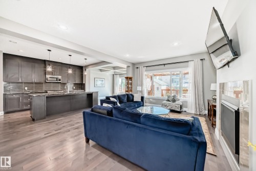 Open-concept living space featuring wood-finish flooring, a contemporary kitchen with dark cabinetry and pendant lighting, a recessed fireplace with a tiled surround, and large windows - 4155 Charles Link, Edmonton, AB - Indoor With Fireplace
