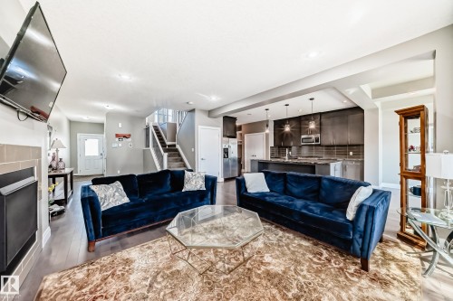 Open-concept living space featuring wood-finish flooring, a modern fireplace with a tiled surround, a contemporary kitchen with dark cabinetry and stainless steel appliances, and a staircase with open risers and metal railings - 4155 Charles Link, Edmonton, AB - Indoor Photo Showing Living Room With Fireplace