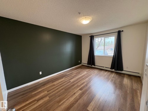 Spacious room featuring wood-finish flooring, a prominent accent wall, and a large window - 138 9525 162 Avenue, Edmonton, AB 