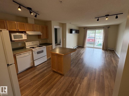 Open-concept living space featuring wood-finish flooring, a kitchen with natural wood cabinetry, a central kitchen island with storage, a built-in microwave, and track lighting - 138 9525 162 Avenue, Edmonton, AB 