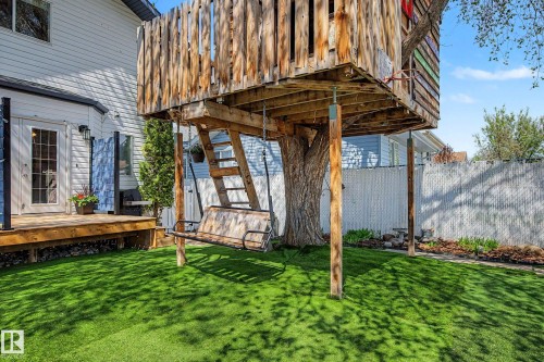 Backyard featuring a custom treehouse structure with a built-in swing, a wood-plank deck with black railings, a white siding exterior, and a manicured green lawn - 16211 55A Street, Edmonton, AB - Outdoor With Deck Patio Veranda