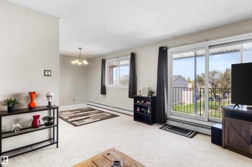 Living area featuring light-colored carpeting, a wall-mounted chandelier, and a sliding glass door providing access to an exterior balcony - 6 3803 76 Street, Edmonton, AB - Indoor