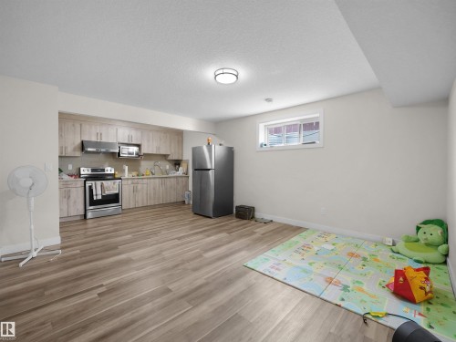 Open-concept living space with wood-finish flooring, a full kitchen featuring light wood-tone cabinetry, stainless steel appliances, and a window - 3247 15 Avenue Nw, Edmonton, AB 