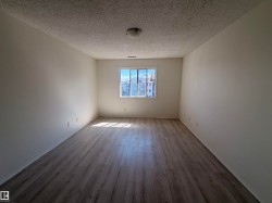 Bright interior space featuring a window with exterior residential structures visible, wood-finish flooring, white walls, and a textured ceiling with a flush-mount light fixture - 