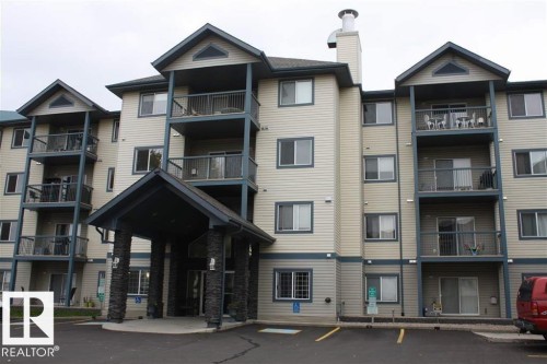 Multi-story residential building featuring light-toned siding with contrasting dark trim, multiple private balconies with metal railings, a covered entrance supported by stone-clad columns, and an integrated chimney structure - 424 16303 95 Street, Edmonton, AB - Outdoor With Balcony With Facade