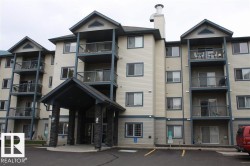 Multi-story residential building featuring light-toned siding with contrasting dark trim, multiple private balconies with metal railings, a covered entrance supported by stone-clad columns, and an integrated chimney structure - 