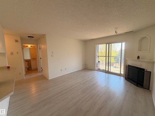 Spacious living area featuring wood-finish flooring, a white mantel fireplace with a black screen, and a sliding glass door leading to an outdoor balcony - 424 16303 95 Street, Edmonton, AB - Indoor Photo Showing Living Room With Fireplace