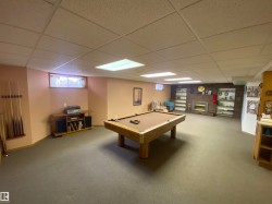 Spacious recreation room featuring a drop-panel ceiling with integrated lighting, wall-to-wall carpeting, and a brick accent wall with a built-in fireplace and shelving - 