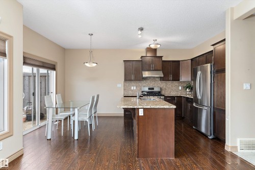 Kitchen and dining area featuring wood-finish flooring, dark wood cabinetry, a central island with a light-toned countertop, and stainless steel appliances including a refrigerator and range - 23 3103 Hilton Drive, Edmonton, AB - Indoor Photo Showing Kitchen With Upgraded Kitchen