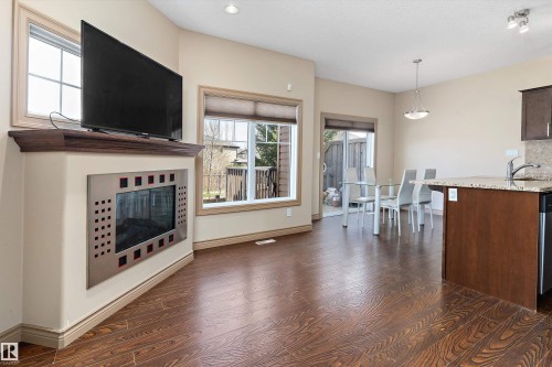 Open-concept living area featuring rich wood-finish flooring, a modern wall-mounted fireplace with a decorative metal surround, and a coordinating wood mantel - 23 3103 Hilton Drive, Edmonton, AB - Indoor