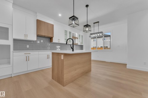Bright kitchen space featuring a wood-finish island with a white countertop, white cabinetry with gold-toned hardware, and a gray subway tile backsplash - 6226 45 Avenue, Beaumont, AB - Indoor Photo Showing Kitchen