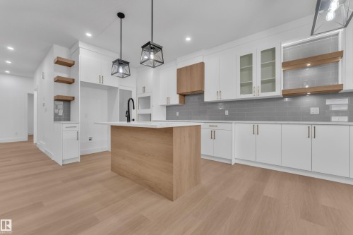 Modern kitchen featuring a central island with wood-finish paneling and a light-toned countertop, white cabinetry with gold-toned hardware, a gray subway tile backsplash, and light wood-finish flooring - 6226 45 Avenue, Beaumont, AB - Indoor Photo Showing Kitchen