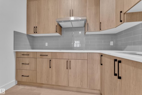 Modern kitchen cabinetry with wood-finish doors and matte black hardware, featuring white countertops, a gray subway tile backsplash, and a stainless steel range hood - 6226 45 Avenue, Beaumont, AB - Indoor Photo Showing Kitchen