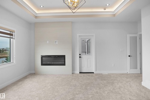 Spacious room featuring a tray ceiling with recessed lighting, a modern electric fireplace with a light-toned surround, light-colored carpeting, and a window offering a water view - 162 Edgewater Circle, Leduc, AB - Indoor With Fireplace
