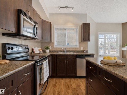 The kitchen features dark wood cabinetry, granite-style countertops, and a full suite of stainless steel appliances including a microwave, oven, and dishwasher - 120 8931 156 Street, Edmonton, AB - Indoor Photo Showing Kitchen With Stainless Steel Kitchen