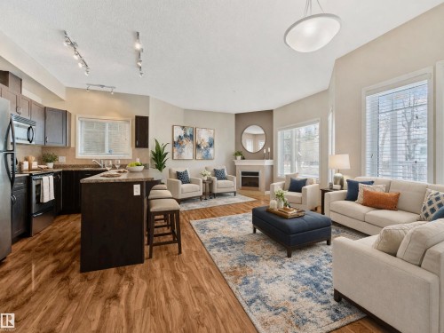 Open concept living space featuring wood-look flooring throughout, a kitchen island with seating, a fireplace with a mantel, and large windows providing natural light - 120 8931 156 Street, Edmonton, AB - Indoor With Fireplace