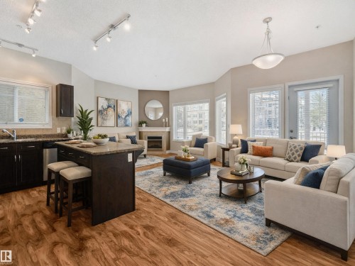 The living area features wood-look flooring, a kitchen island with a stone countertop, and a fireplace with a stone surround - 120 8931 156 Street, Edmonton, AB - Indoor Photo Showing Living Room With Fireplace