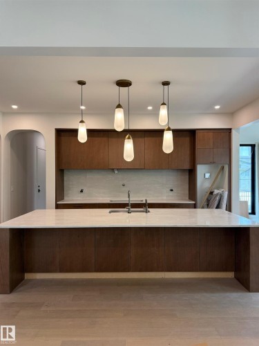 Modern kitchen featuring a large island with a light-toned countertop, rich wood-finish cabinetry, a built-in sink with a brushed metal faucet, a light-toned backsplash, and contemporary pendant lighting - 14702 Park Drive, Edmonton, AB - Indoor Photo Showing Kitchen With Double Sink