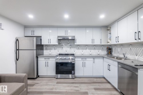 Modern kitchen featuring stainless steel appliances, white flat-panel cabinetry with black pulls, gray countertops, and a striking geometric tile backsplash - 6607 50A Avenue, Beaumont, AB - Indoor Photo Showing Kitchen With Stainless Steel Kitchen