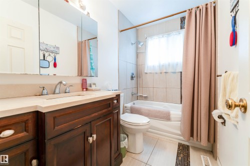 Bathroom featuring a rich wood-finish vanity with an integrated sink and light-toned countertop, a mirrored medicine cabinet, a tiled shower-tub combination, and tiled flooring with an accent strip - 13231 74 Street, Edmonton, AB - Indoor Photo Showing Bathroom