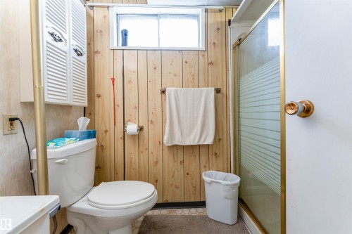 Bathroom featuring wood paneling, a white toilet, a shower enclosure with a patterned glass door, a wall-mounted towel bar, and a louvered wall cabinet - 13231 74 Street, Edmonton, AB - Indoor Photo Showing Bathroom