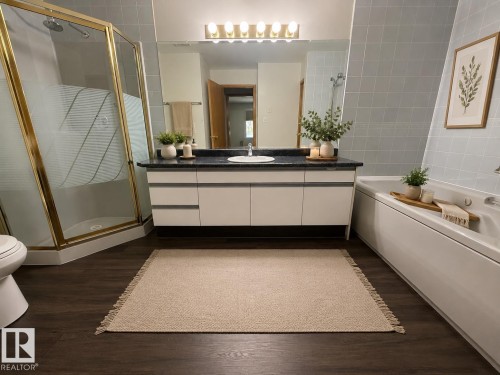 Bathroom featuring a dark wood-finish floor, a gold-framed glass shower enclosure, a vanity with white cabinetry and dark countertop, a mirror with sconce lighting, and a white soaking tub with tiled surround - 309 10610 76 Street, Edmonton, AB - Indoor Photo Showing Bathroom