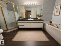 Bathroom featuring a dark wood-finish floor, a gold-framed glass shower enclosure, a vanity with white cabinetry and dark countertop, a mirror with sconce lighting, and a white soaking tub with tiled surround - 