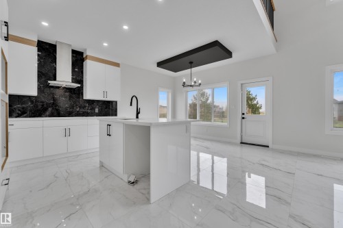 Modern kitchen and living area featuring a large central island, white cabinetry with wood accents, a black slab backsplash, stainless steel range hood, and polished tile flooring throughout - 12623 44 Street, Edmonton, AB 