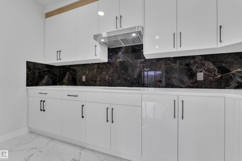 Modern kitchen featuring white cabinetry with matte black hardware, a stainless steel range hood, and a dark marble backsplash - 12623 44 Street, Edmonton, AB 