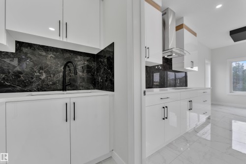 Modern kitchen featuring a sleek black backsplash, white cabinetry with contemporary hardware, and a stainless steel range hood - 12623 44 Street, Edmonton, AB 