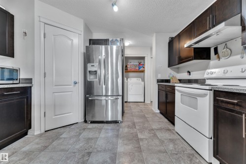 Kitchen with appliances with stainless steel finishes, dark brown cabinetry, under cabinet range hood, a textured ceiling, and washer and dryer - 6152 13 Avenue, Edmonton, AB - Indoor Photo Showing Kitchen