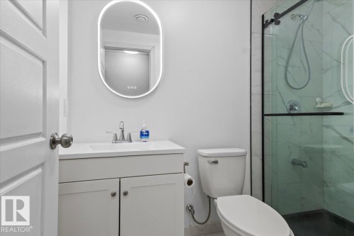 Modern bathroom featuring a contemporary vanity with an integrated sink, an illuminated oval mirror, a commode, and a glass-enclosed shower with white marble-patterned tile - 236 39 Avenue, Edmonton, AB - Indoor Photo Showing Bathroom