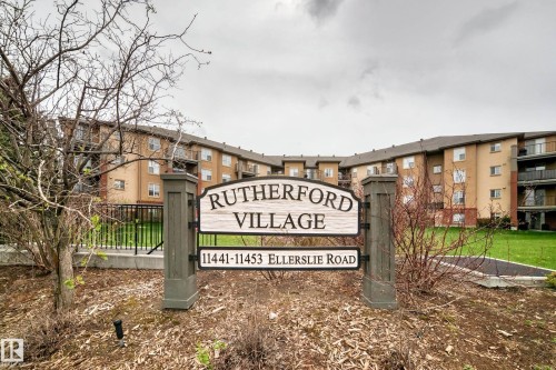 "Rutherford Village" entry signage featuring a multi-story residential building with brick and stucco exterior, multiple windows, and individual balconies - 113 11441 Ellerslie Road, Edmonton, AB - Outdoor