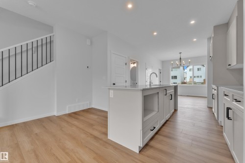 Open-concept kitchen featuring a central island with an undermount sink and contemporary faucet, light-toned wood-finish flooring, white cabinetry with black hardware, recessed lighting, and a modern chandelier - 17330 6A Street Ne, Edmonton, AB - Indoor Photo Showing Kitchen