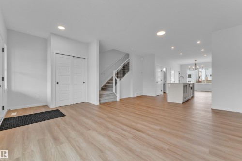 Open-concept living area featuring light wood-finish flooring, recessed lighting, a central staircase with carpeted treads and white risers, and a kitchen island with a built-in sink and seating overhang - 17330 6A Street Ne, Edmonton, AB - Indoor