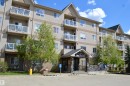 Multi-story residential building featuring a stone-clad entrance with an overhang, multiple balconies with glass railings, a pitched roofline, and mature landscaping - 210 5005 165 Avenue, Edmonton, AB  - Outdoor With Balcony With Facade 