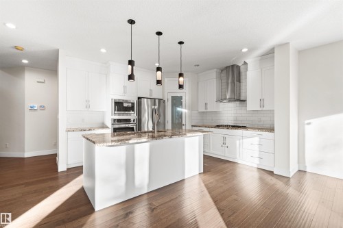 Open-concept kitchen featuring an oversized island with pendant lighting, white shaker cabinetry, stainless steel appliances, and wood-finish flooring - 2036 Ainslie Link Link, Edmonton, AB - Indoor Photo Showing Kitchen With Upgraded Kitchen